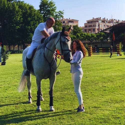 Jinete montado en un caballo blanco siendo asistido por una mujer en un evento ecuestre en un campo verde con edificios al fondo en Burgos.