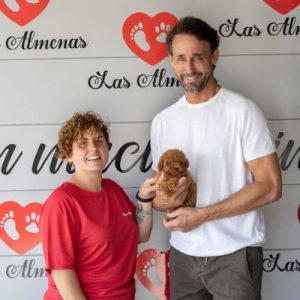 Hombre sosteniendo un cachorro junto a una empleada de la Residencia Canina Las Almenas, ambos sonrientes frente al logotipo de la residencia.